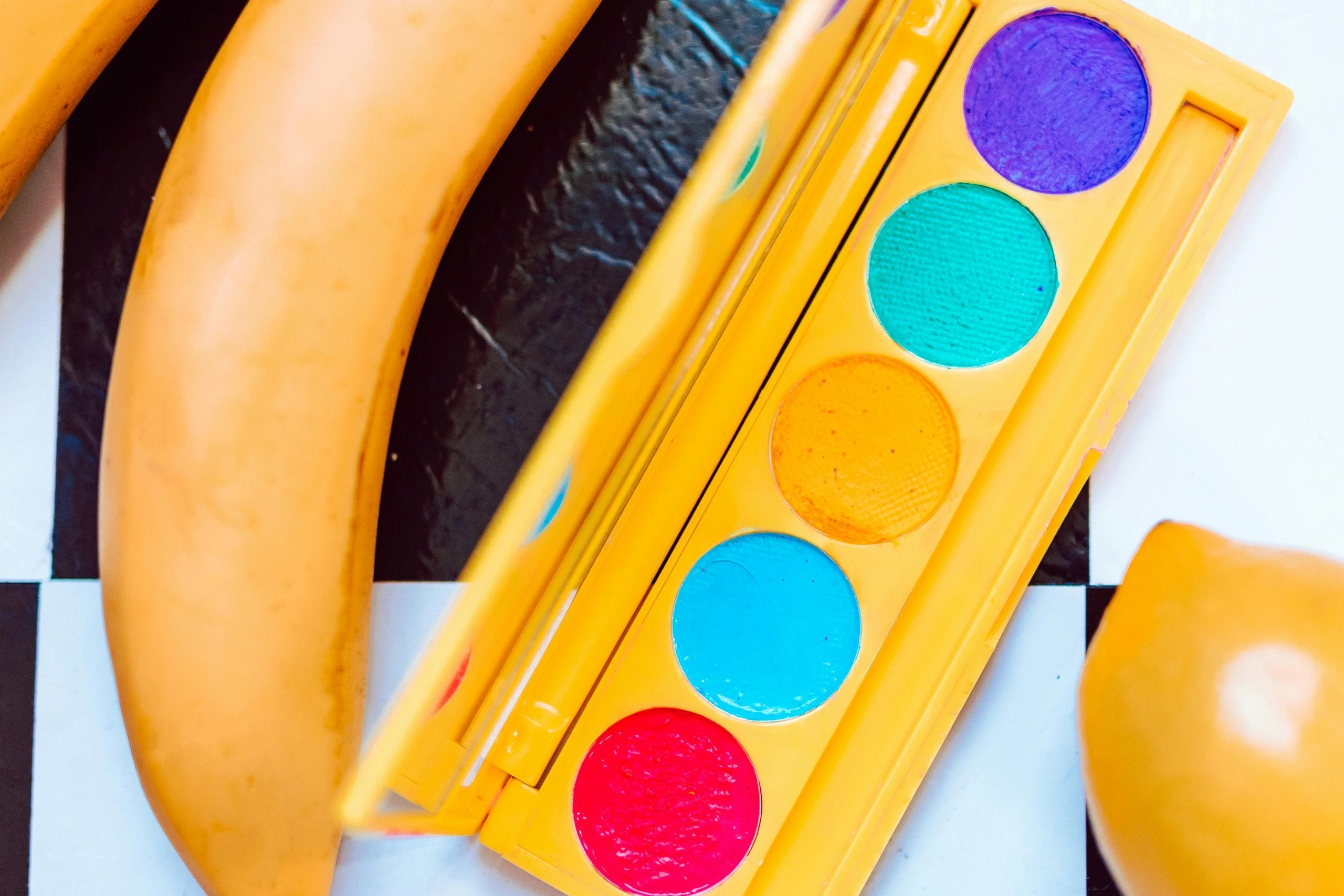 A flat lay of a bright, colorful makeup palette alongside bananas on a checkered surface.